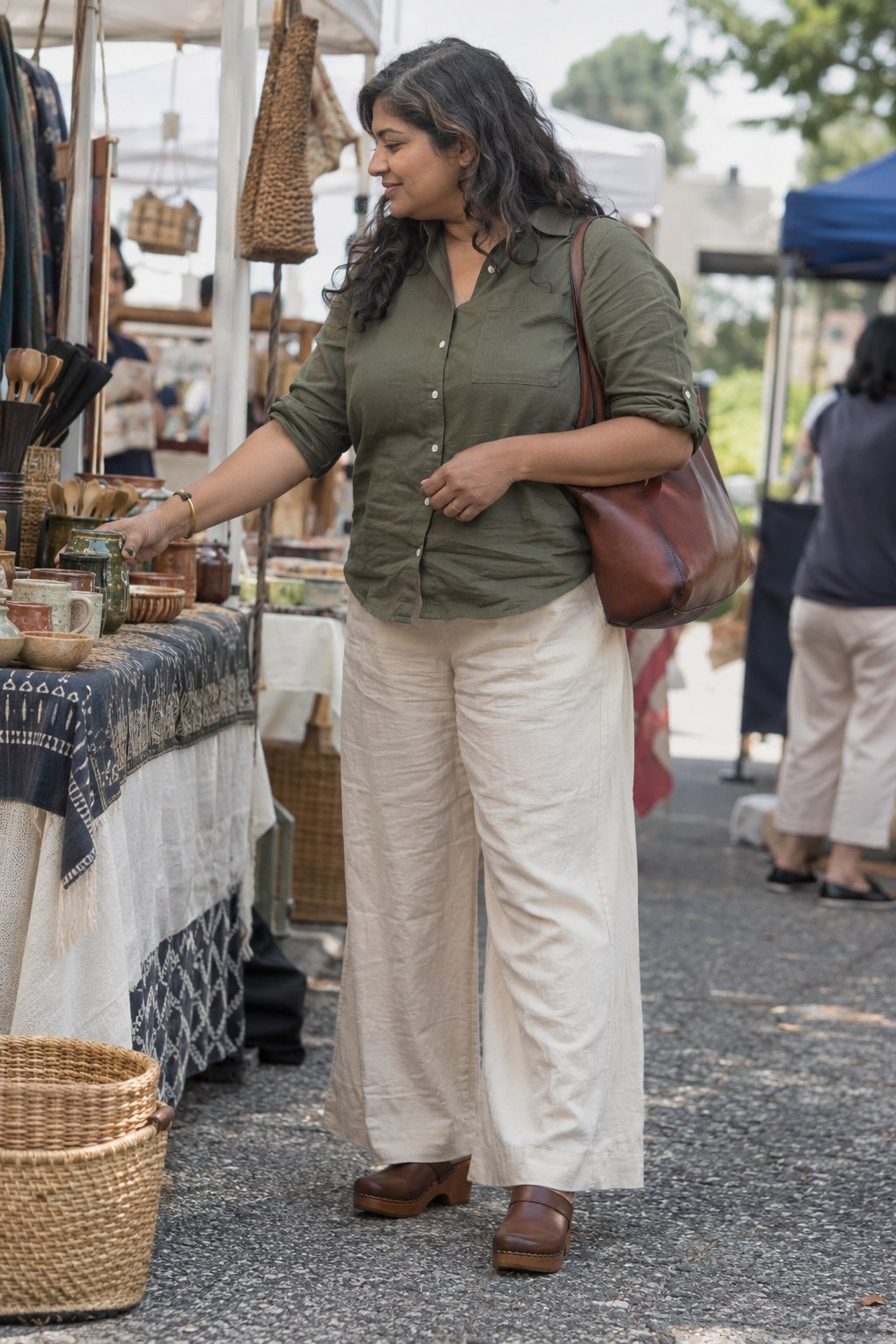 Woman wearing cream linen trousers, an olive cotton shirt and brown chunky clogs at an outdoor market.