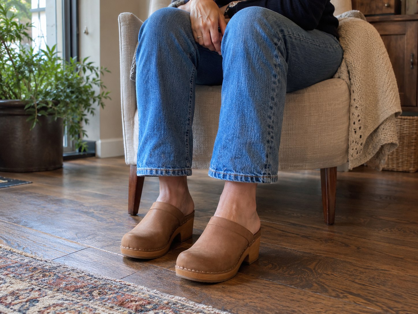 Woman seated at home trying tan chunky clogs with straight jeans, with both shoes resting flat on the floor.