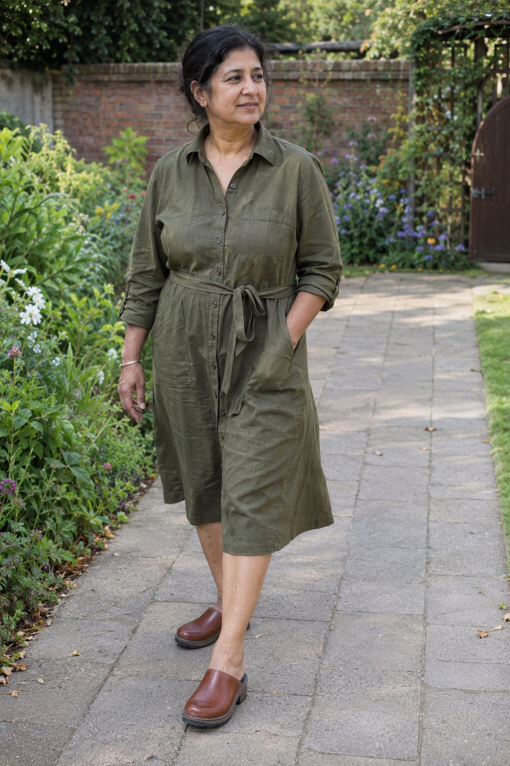 Woman in an olive cotton shirt dress with a tied waist and brown chunky clogs on a garden path.