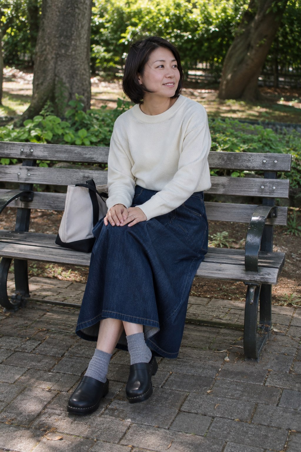 Woman in a denim midi skirt, ivory sweater and black chunky clogs with gray socks.
