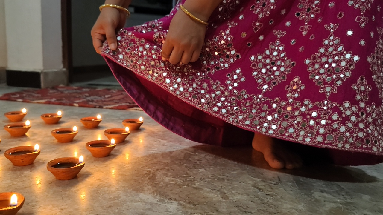 Close-up of hands holding up the edge of a magenta lehenga with mirror work while stepping past rows of lit diyas on the floor