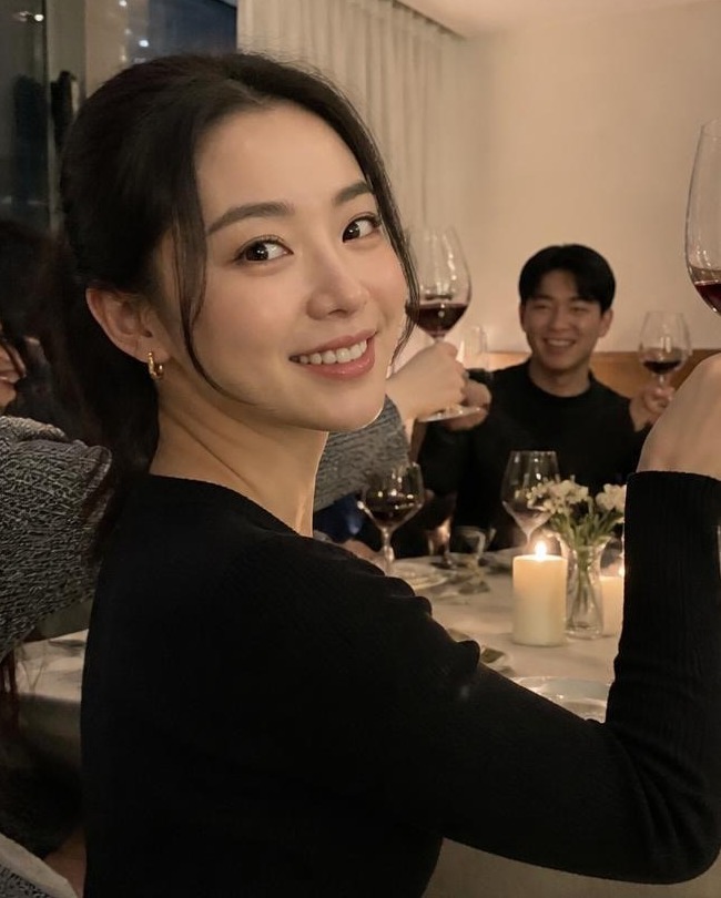 Smiling woman with sleek pulled-back hair, small gold hoop earrings and soft minimal makeup raising a glass at a candlelit dinner