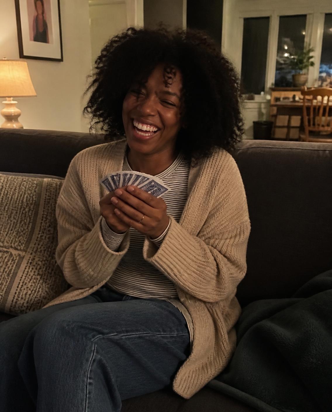 Woman with natural curly hair in an oatmeal cardigan and striped long-sleeve tee laughing on a couch while holding a hand of playing cards close to her chest