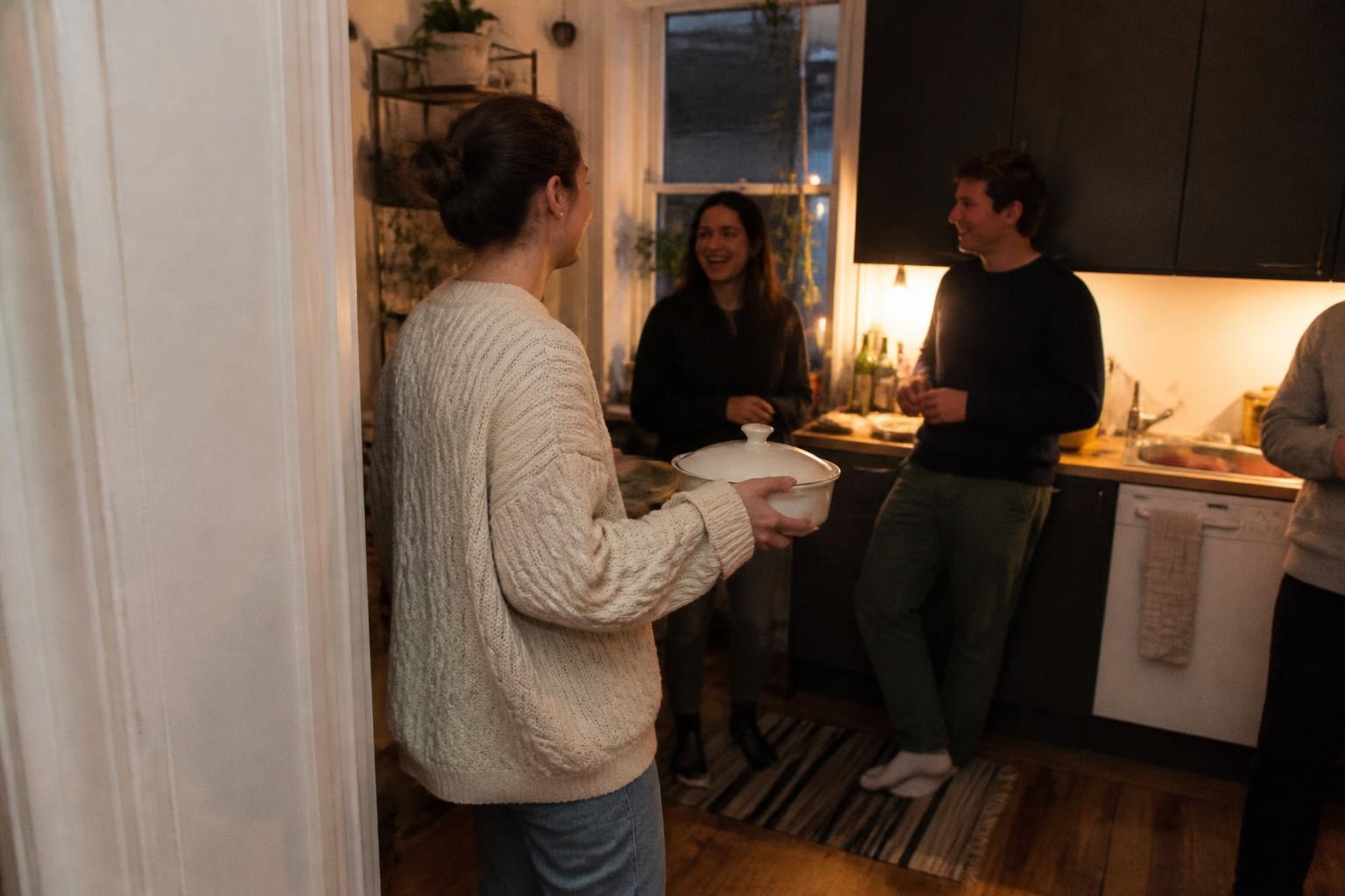 Woman in an oversized cream cable-knit sweater and jeans carrying a covered casserole dish into a small apartment kitchen where friends are chatting