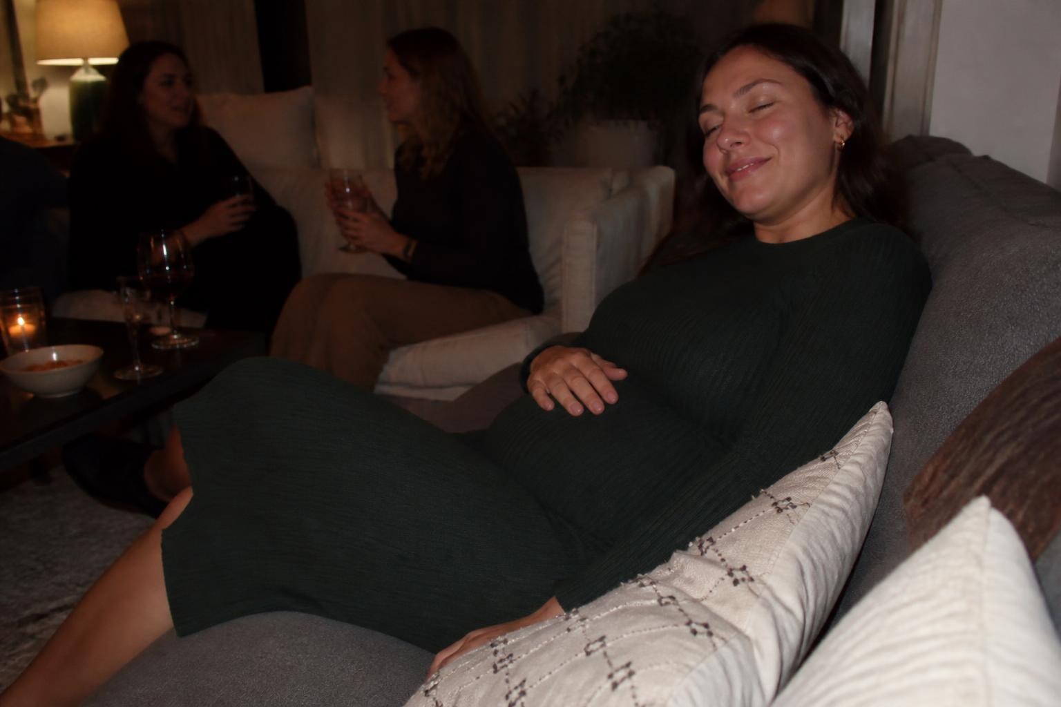 Woman in a dark green ribbed knit midi dress leaning back on a sofa after dinner with her eyes closed and a satisfied smile