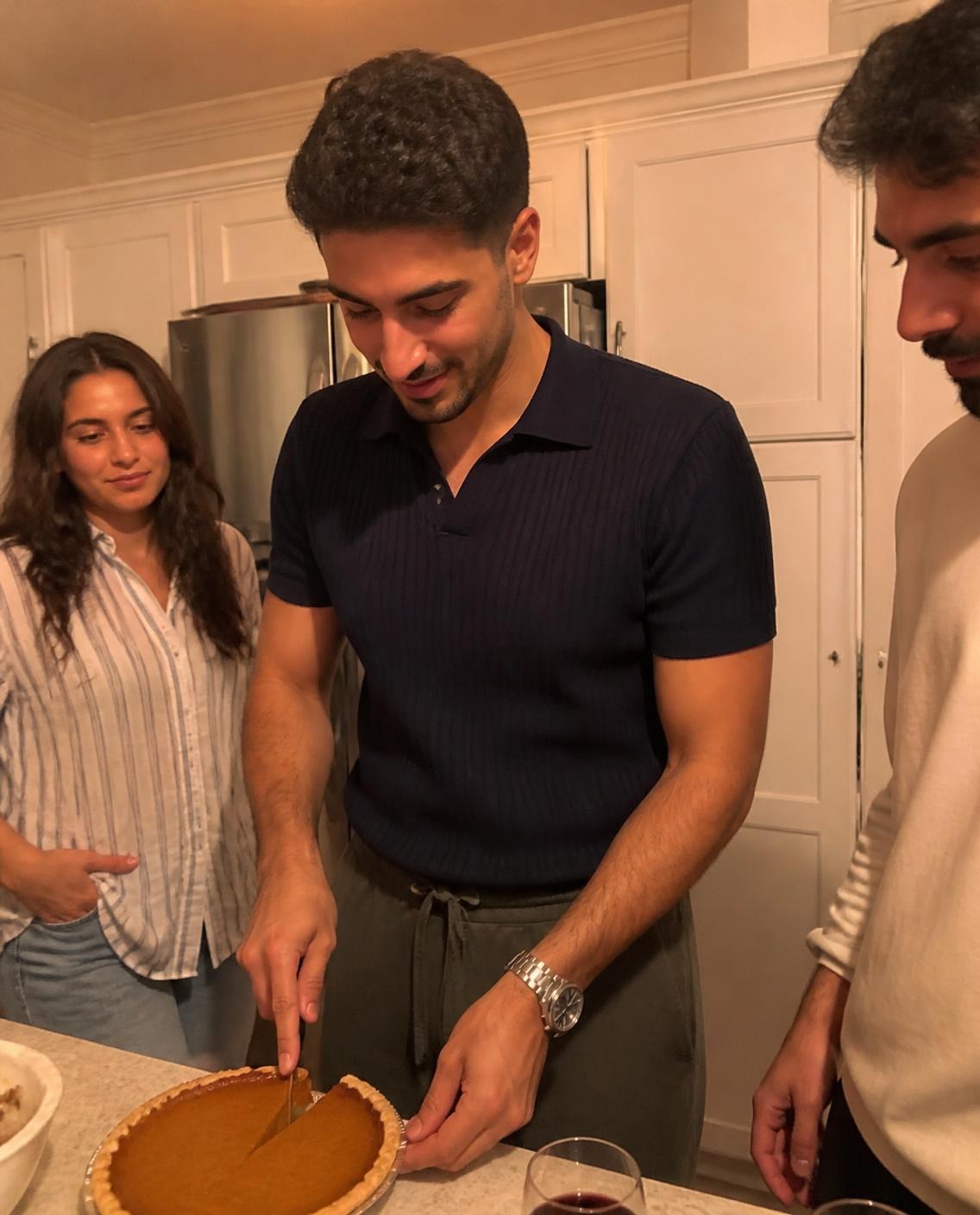 Man in a navy knit polo and relaxed olive drawstring trousers cutting a slice of pumpkin pie in a home kitchen