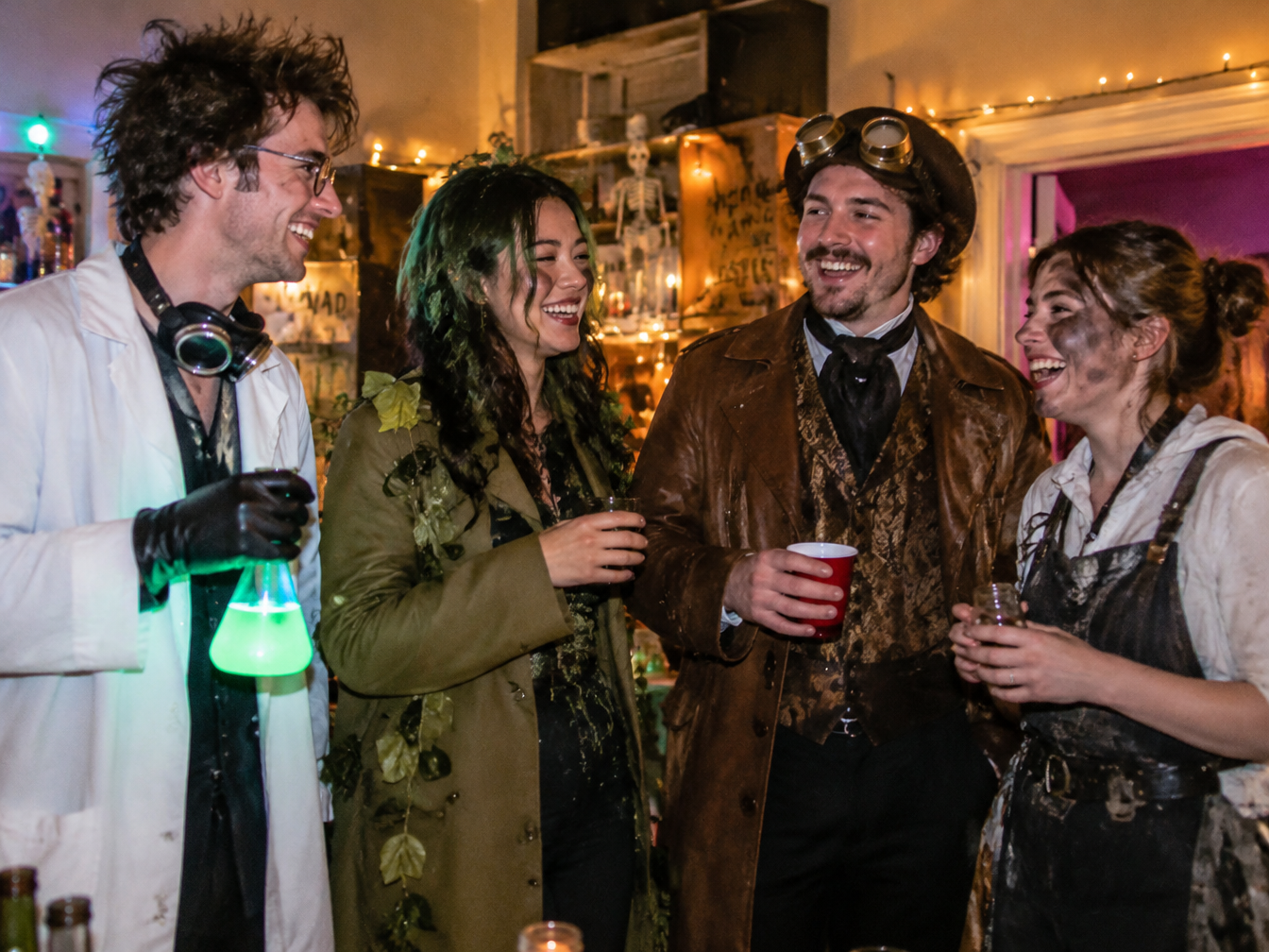 Four friends in different mad scientist costumes laughing together at a Halloween party