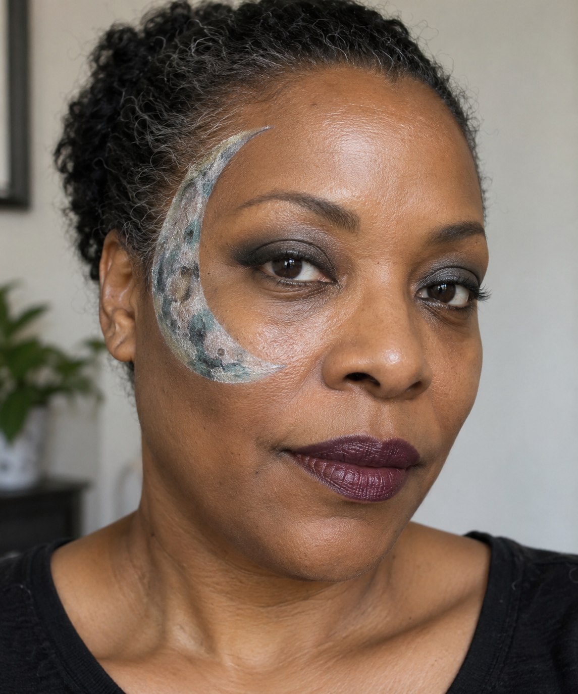 Halloween makeup with a silver crescent on one temple and muted teal shading on the cheek.