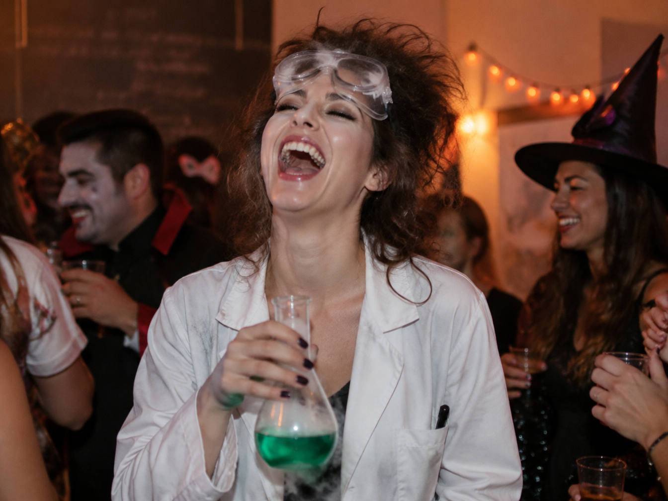 Woman in a white lab coat and goggles laughing with her head thrown back at a Halloween party