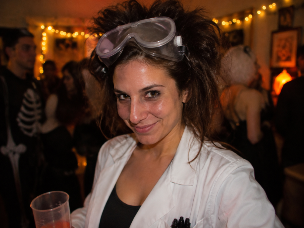Confident woman in a white lab coat with goggles on her forehead and wild hair at a Halloween party