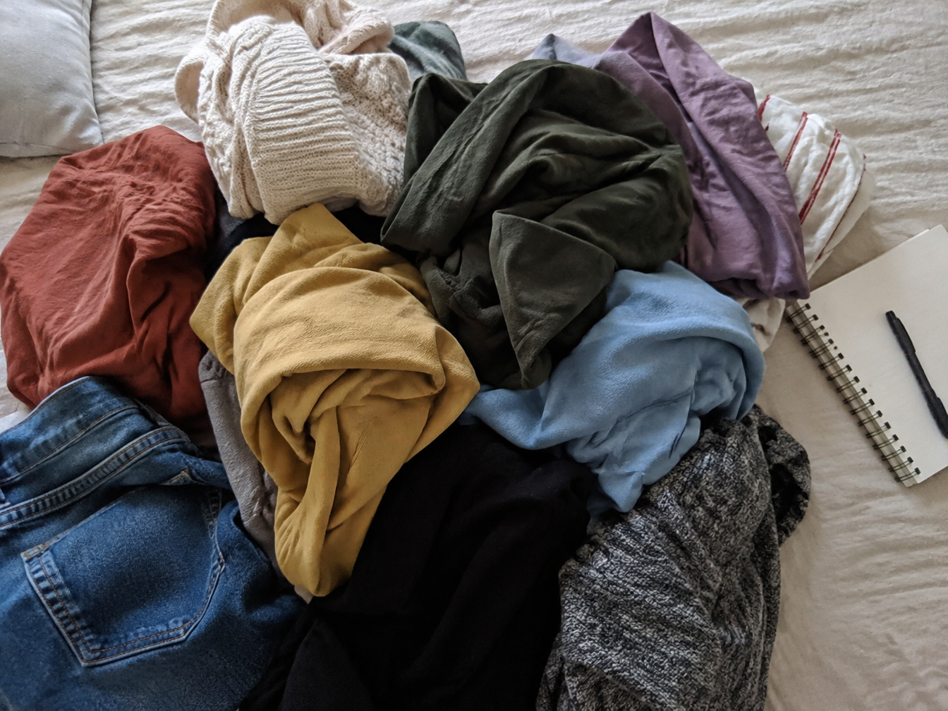 A messy pile of colorful clothes on a bed beside a notebook and pen