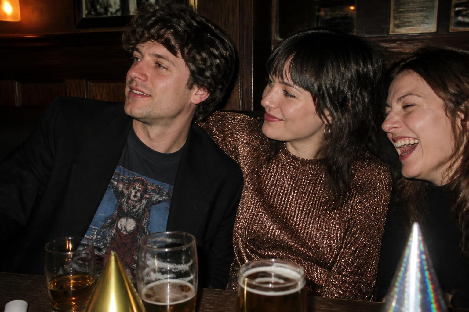 Friends in a wooden bar booth with pints and party hats, one in a black blazer over a band t-shirt, one in a metallic copper knit sweater
