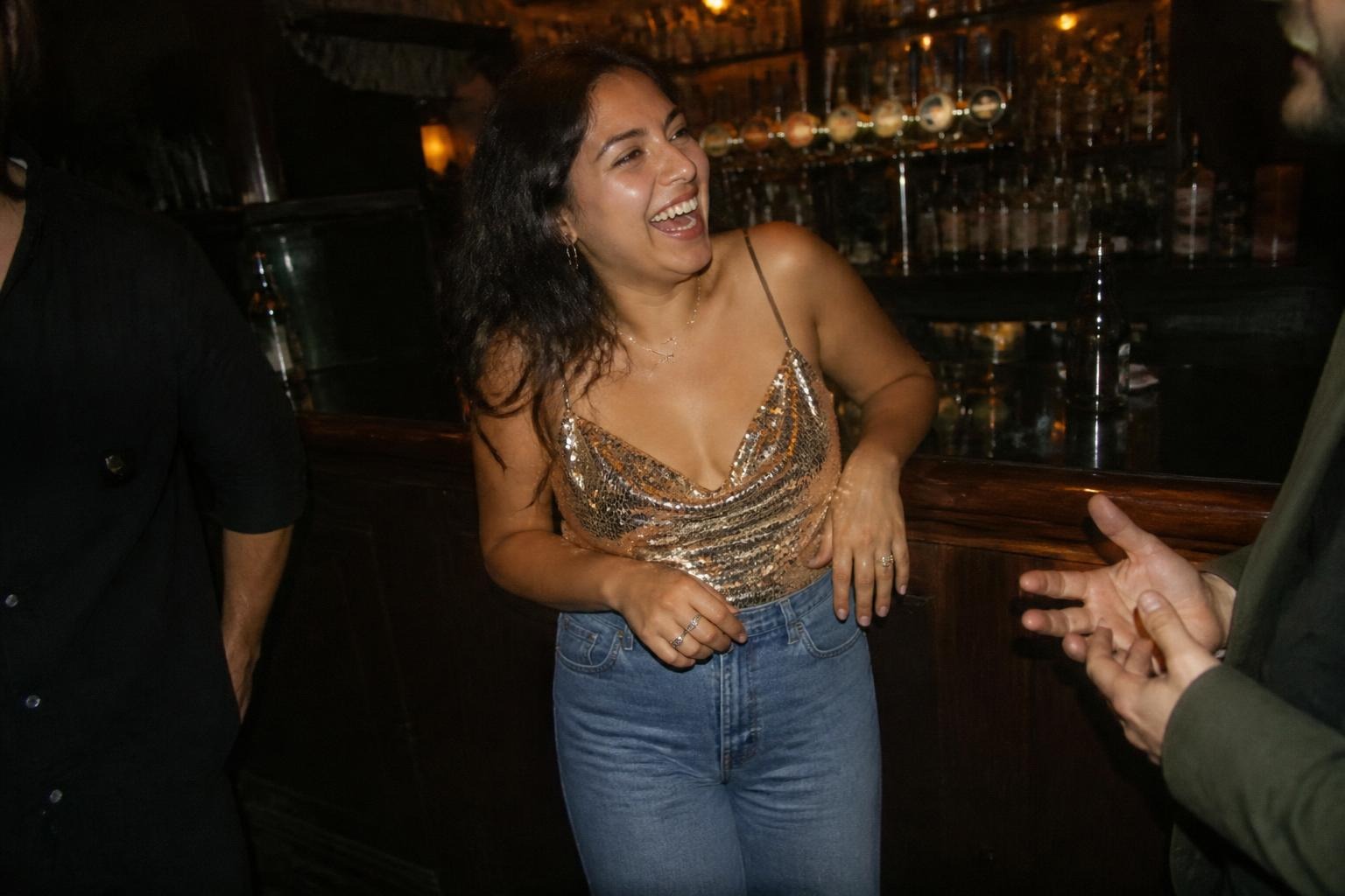 Woman in high-waisted jeans and a gold sequin camisole laughing at a neighborhood bar counter