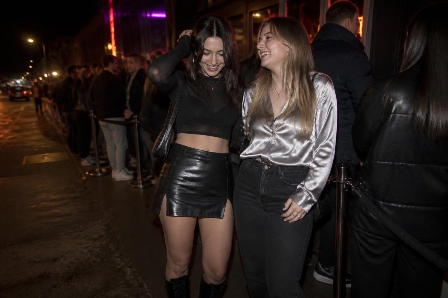 Two friends waiting in line outside a nightclub at night, one in a leather mini skirt and sheer top, the other in a metallic silver shirt