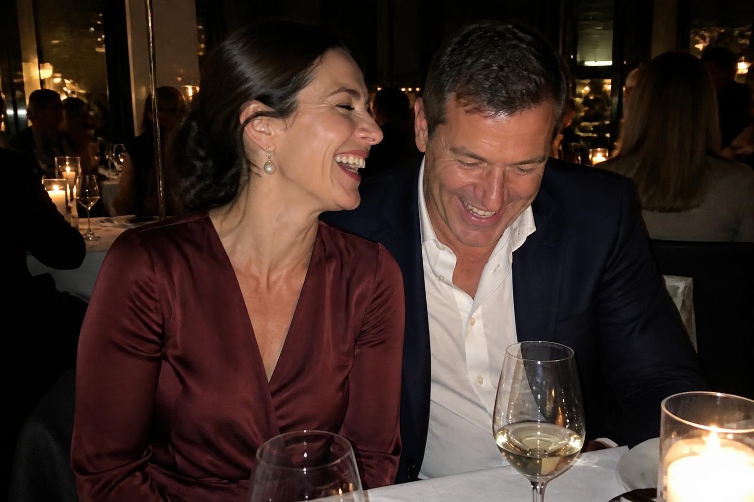 Couple laughing at a candlelit restaurant table, the woman in a burgundy satin wrap dress and pearl earrings, the man in a navy suit
