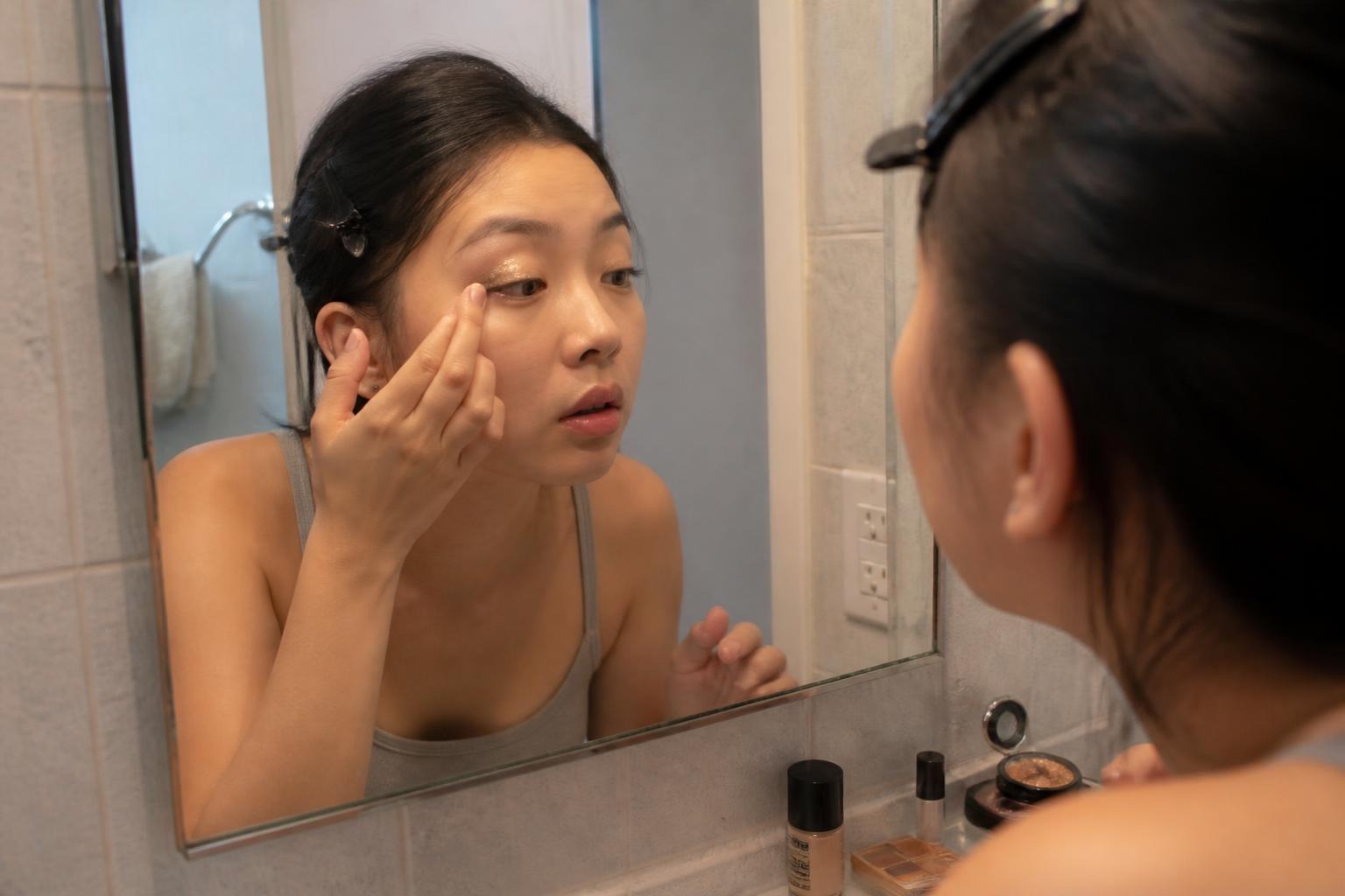 Woman leaning into a bathroom mirror pressing glitter onto her eyelid with her fingertip