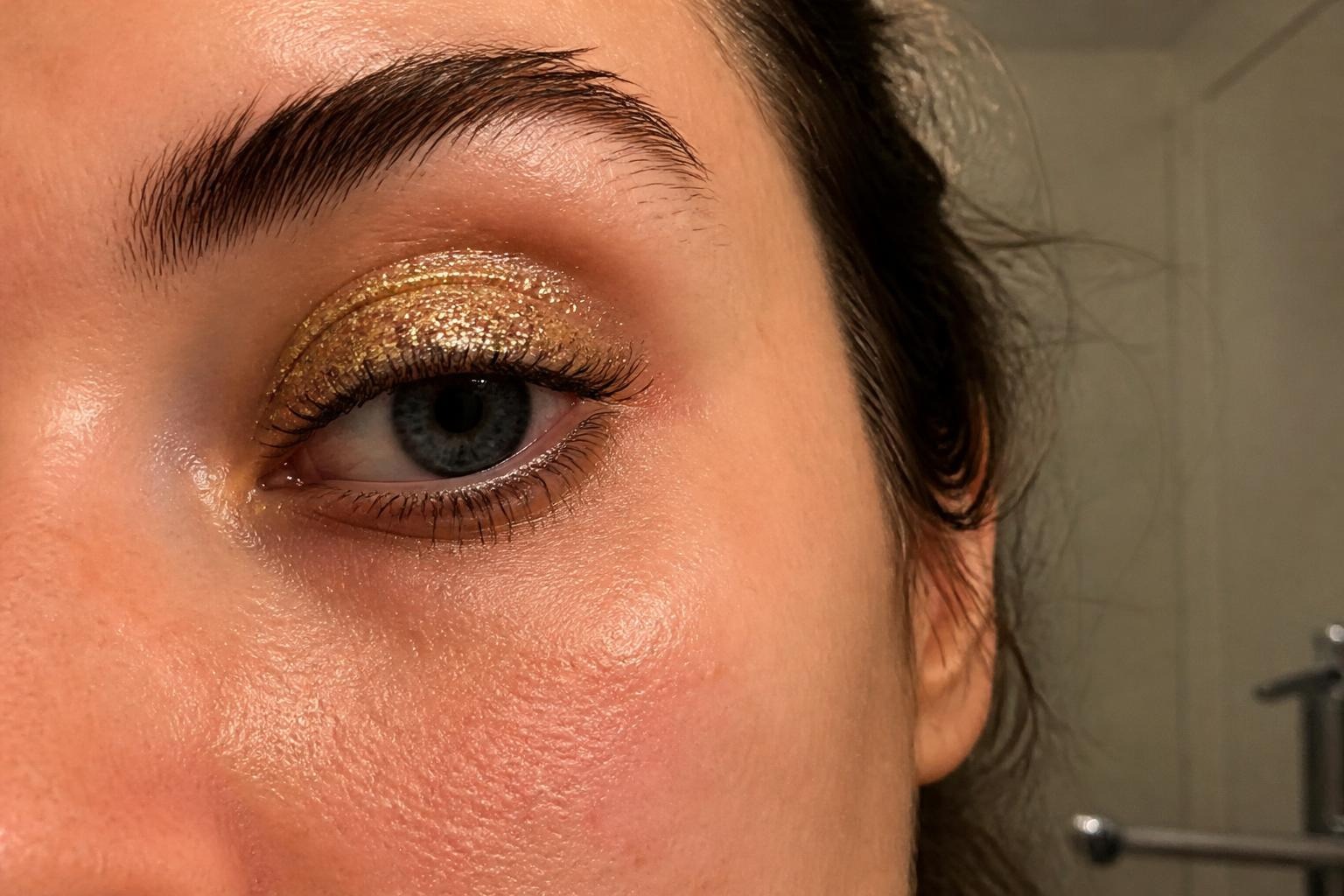 Close-up of a woman's eye with gold glitter eyeshadow on the lid, natural skin texture visible in soft mirror light