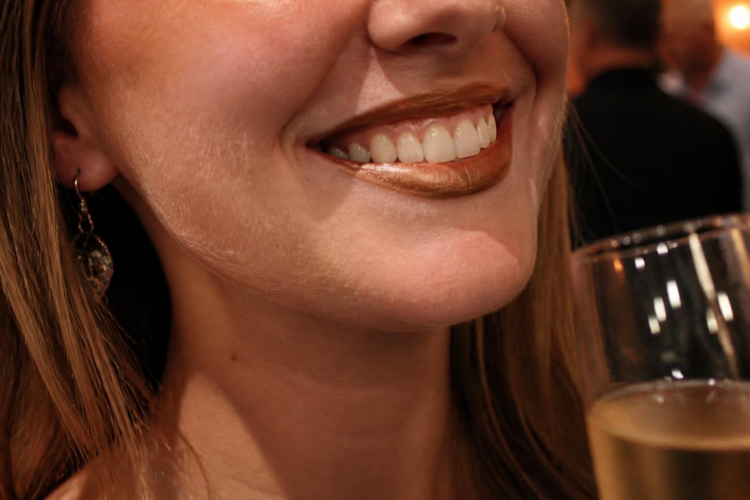 Close-up of a smiling woman wearing metallic bronze lipstick, with a glass of champagne in the foreground