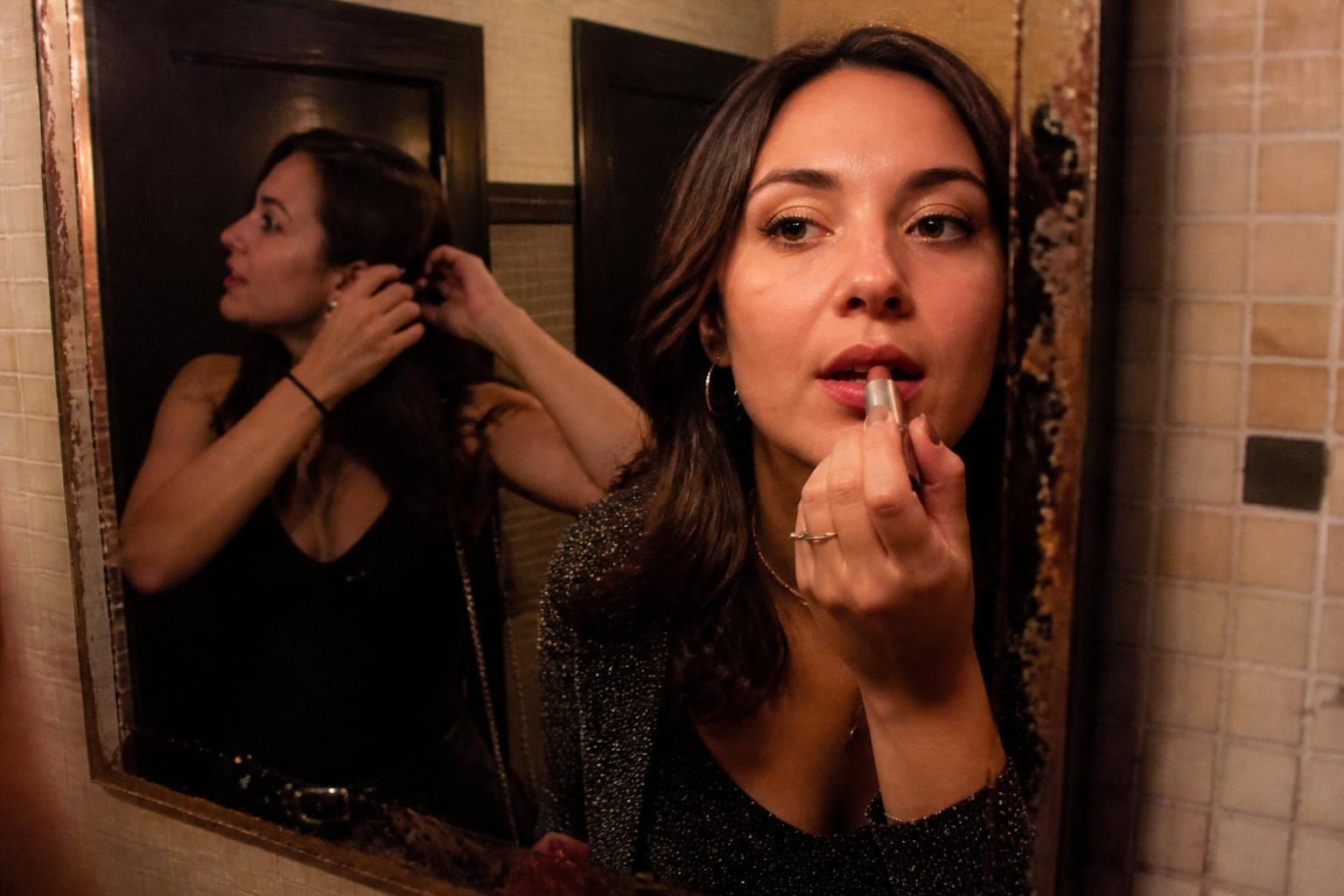 Woman applying lipstick in a tiled bar bathroom mirror while her friend fixes her hair behind her