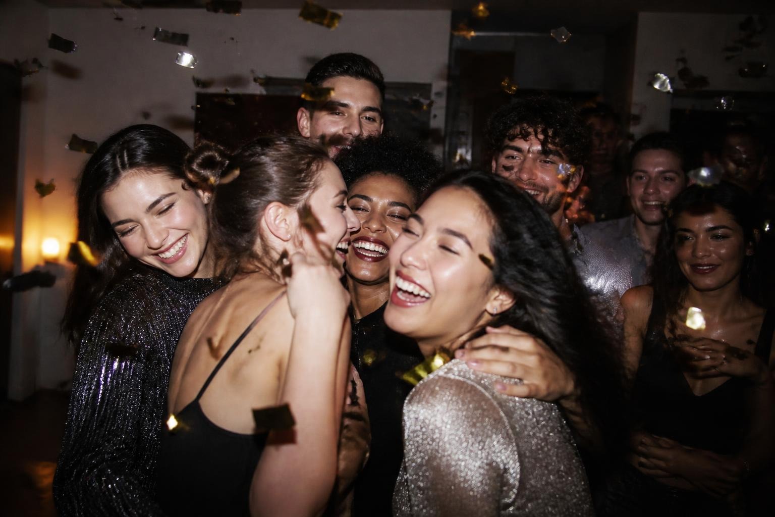 Friends hugging and laughing under falling confetti at midnight at a crowded New Year's Eve party