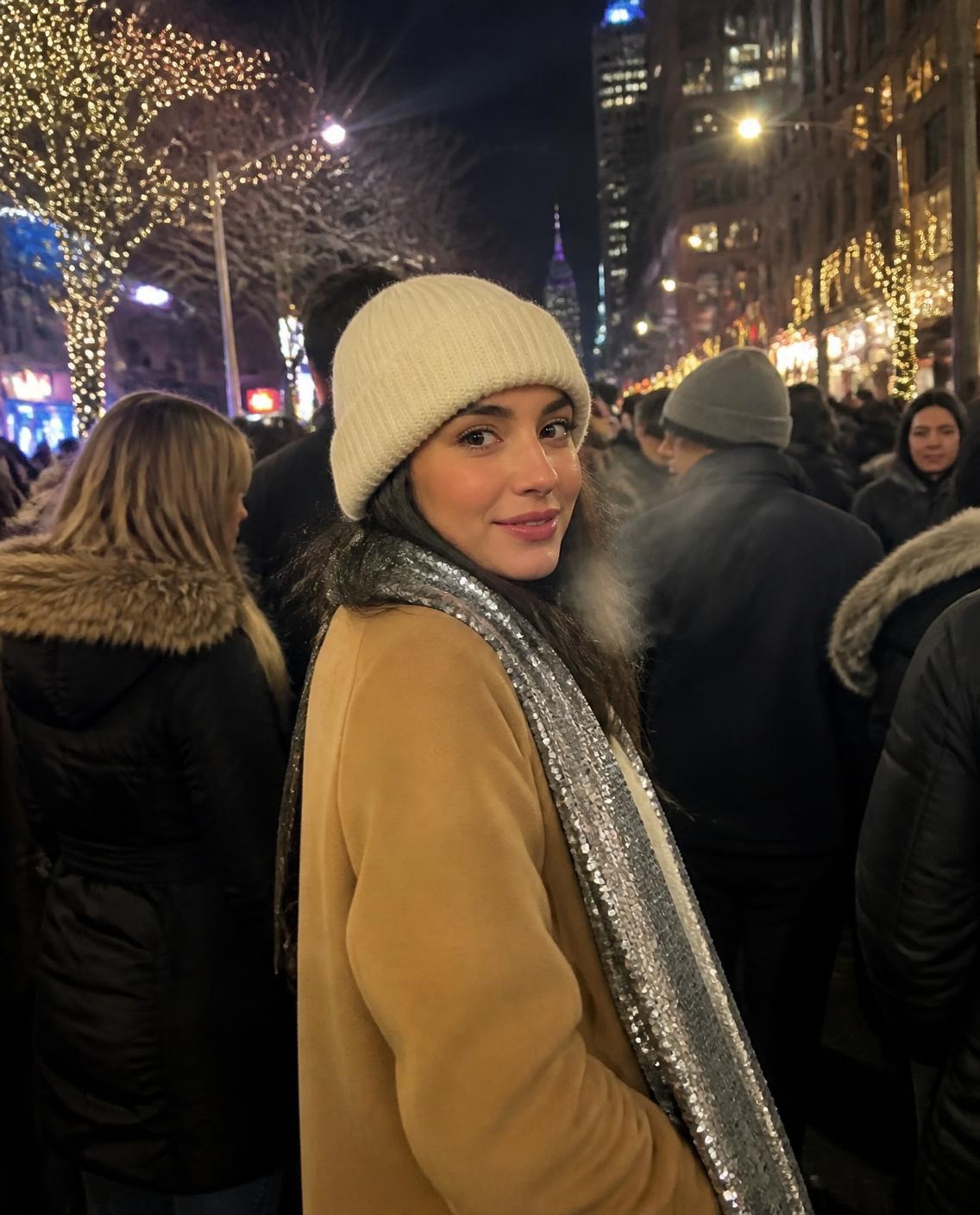 Woman in a camel wool coat, cream beanie and sparkly silver scarf standing in a crowd on a cold city street at night