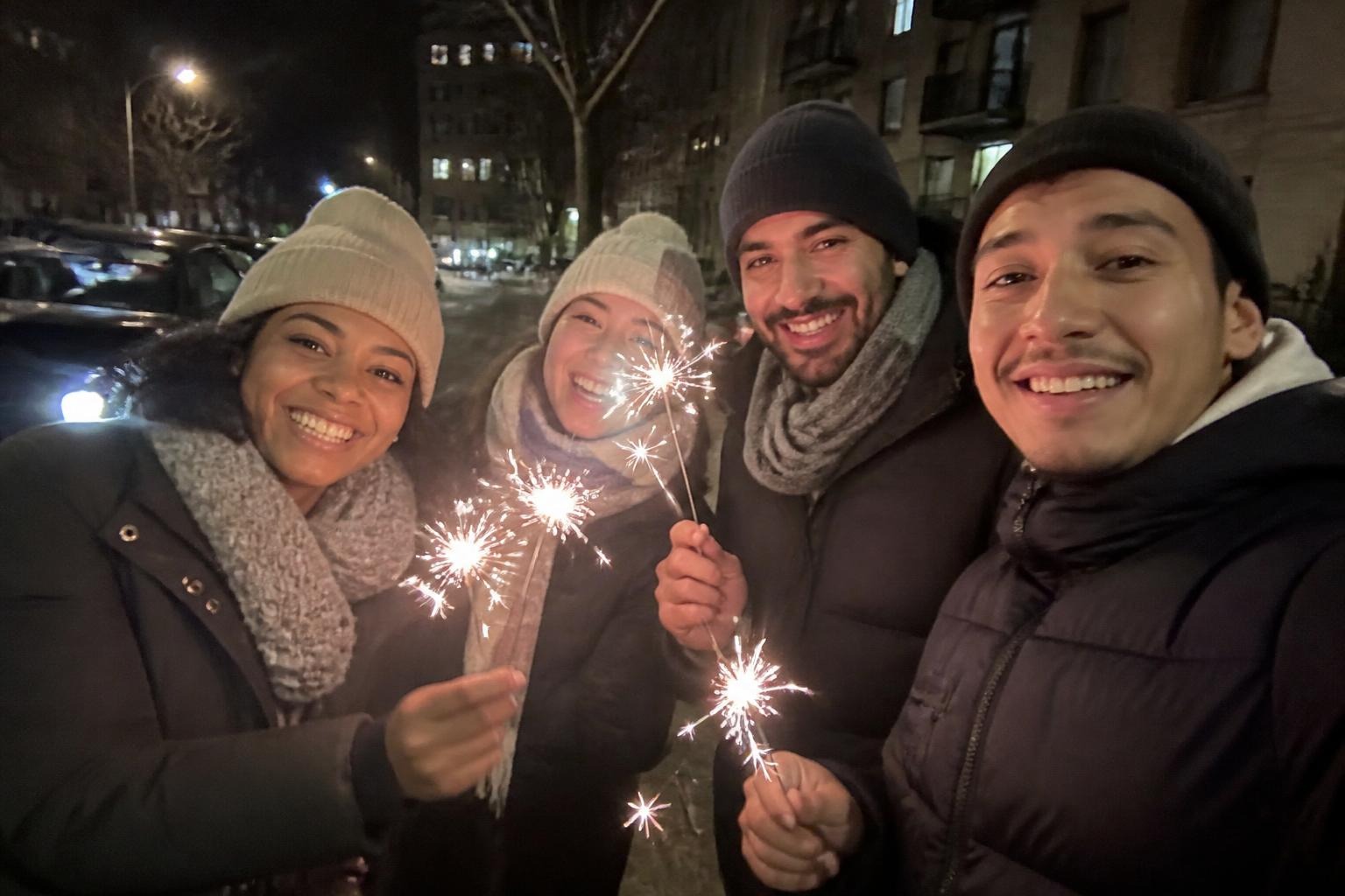 Friends in puffer jackets, scarves and beanies holding lit sparklers on a city street on New Year's Eve