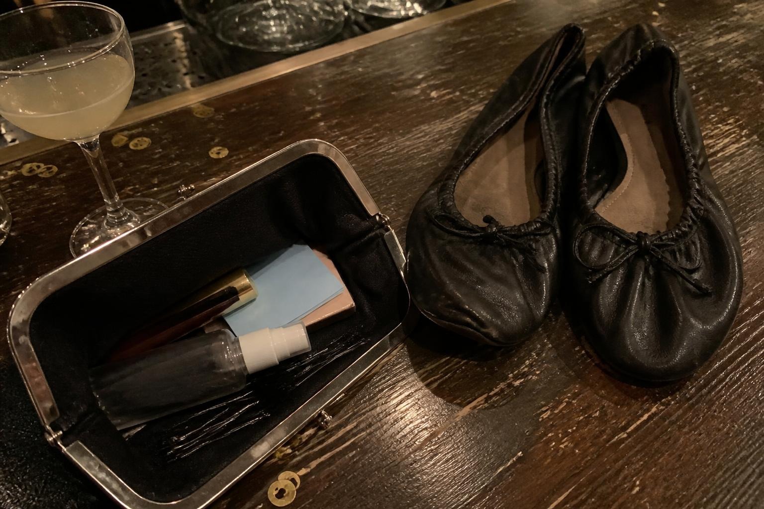 Open metal-frame clutch on a bar table holding lipstick, blotting papers, setting spray and bobby pins, next to folded black ballet flats and a cocktail