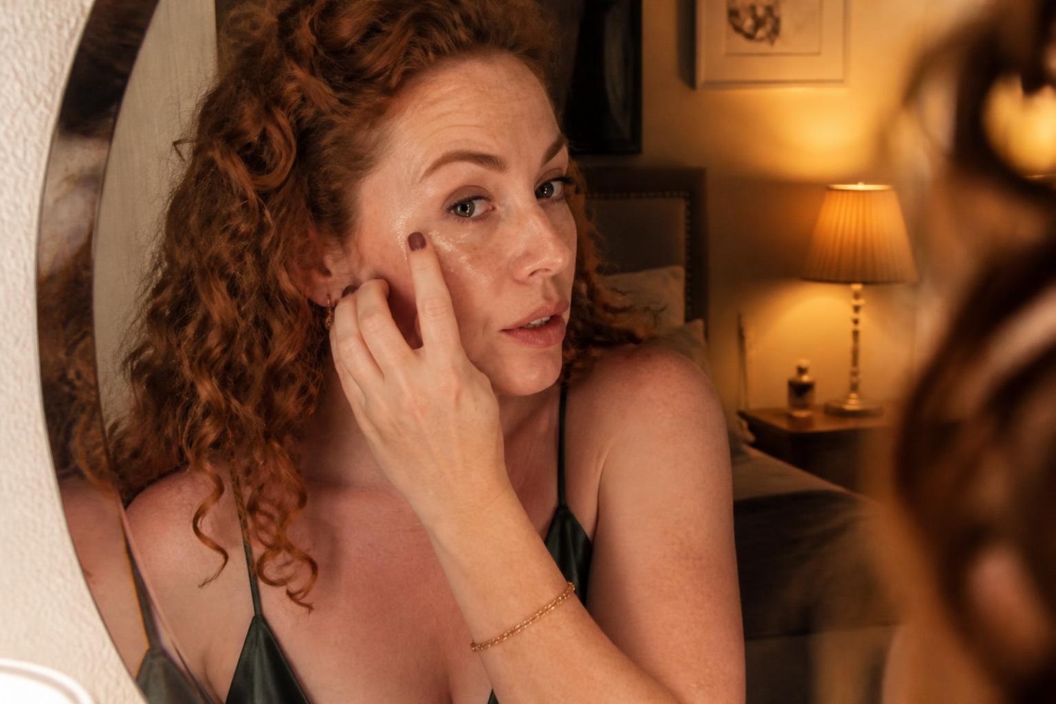 Woman with curly red hair and freckles tapping highlighter onto her cheekbone in a vanity mirror while getting ready
