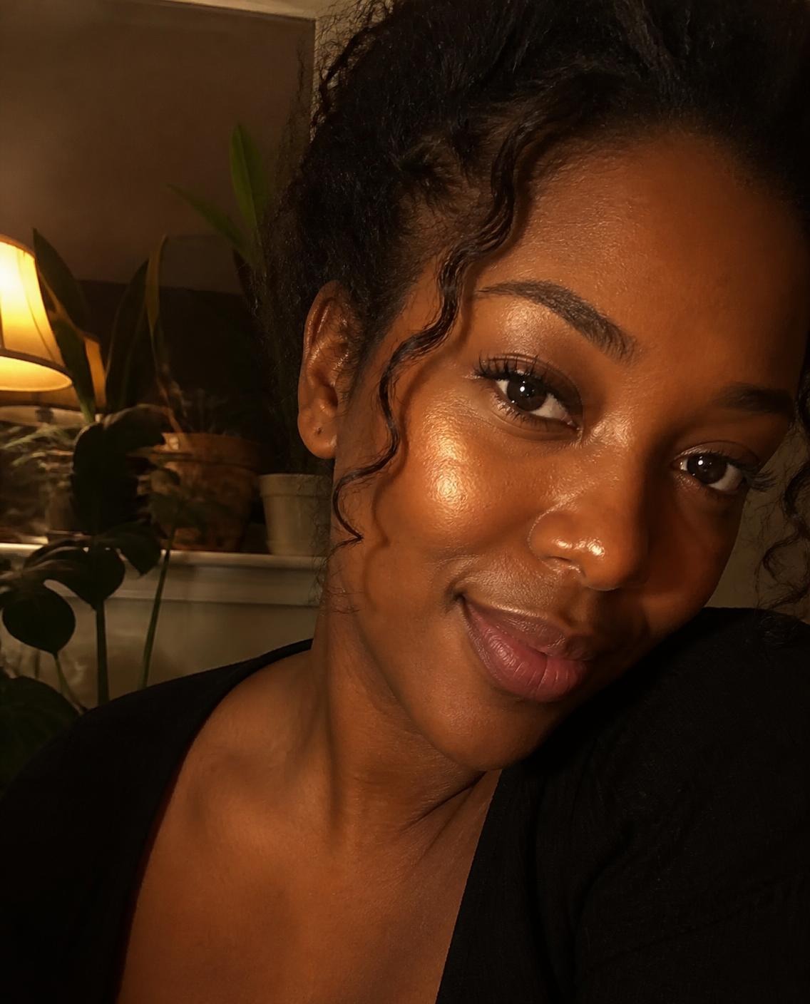 Woman with deep brown skin and a warm golden highlighter glow on her cheekbones and nose, photographed in lamp light