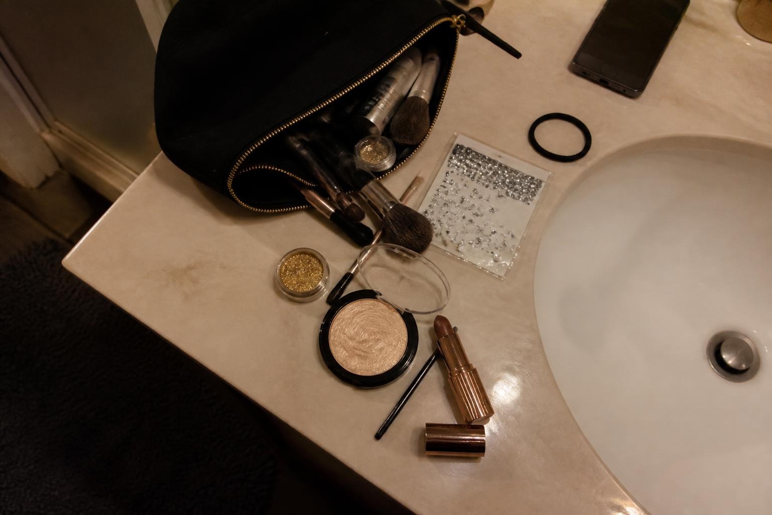 Bathroom counter with an open makeup bag, highlighter compact, lipstick, brushes and a packet of face gems beside the sink