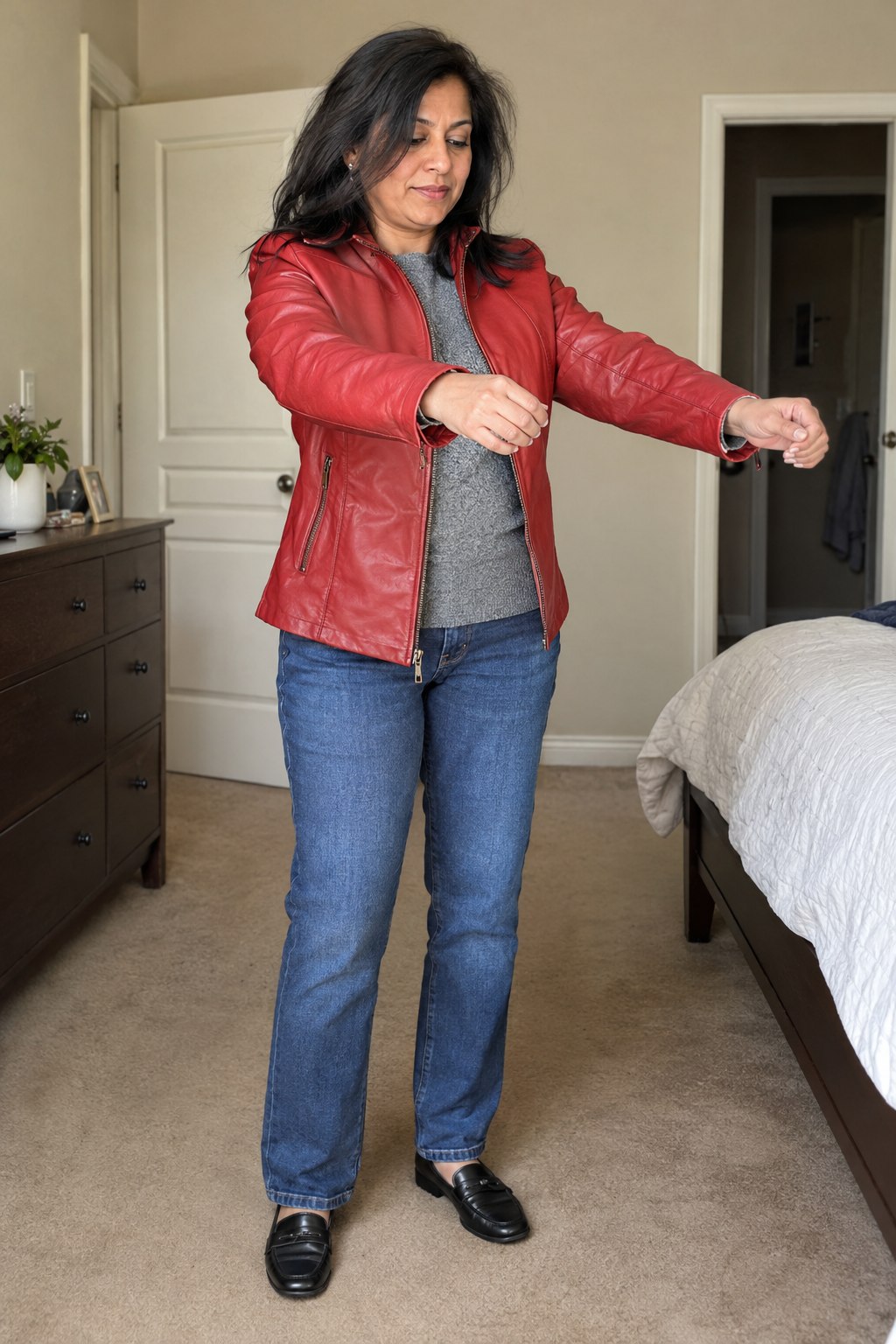 Woman checking the fit of a red leather jacket over a gray knit and jeans at home.